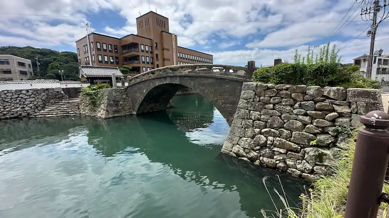 長崎県平戸市の江戸時代に造られた石橋・幸橋。アーチ型の橋が緑がかった川にかかり、背後に近代的な建物が立ち並ぶ風