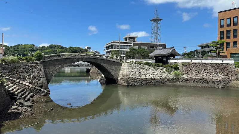 長崎県平戸市にある江戸時代に築造された石造りの幸橋。アーチ型の歴史的石橋が川にかかり、青空の下に門や現代の建物が並ぶ風景