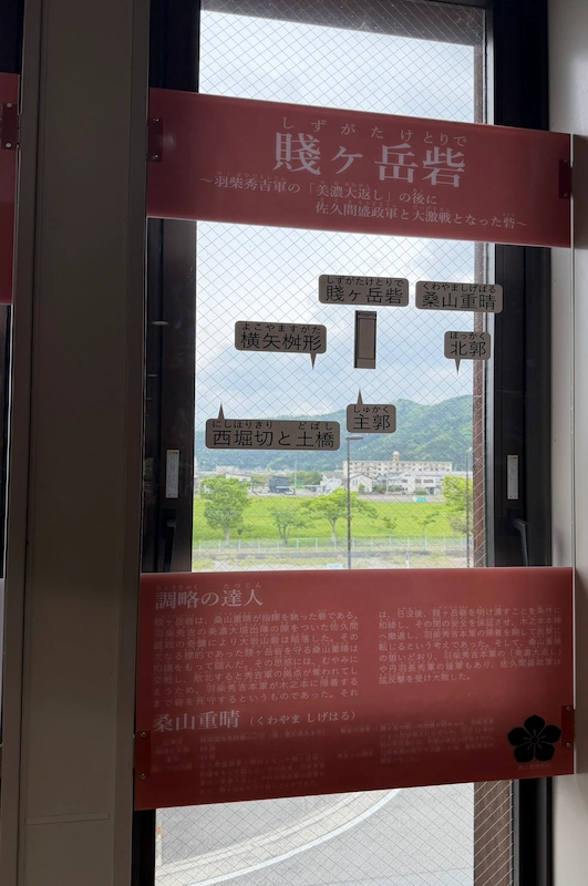 木ノ本駅2階コンコースの窓パネル「賤ヶ岳砦」。窓の外の山並みに砦名の吹き出しが重なる