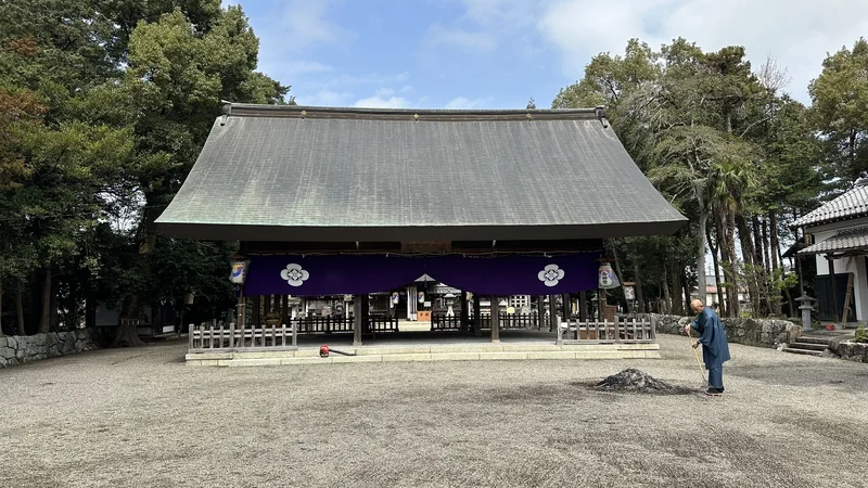 A front view of the worship hall at Ikutsuhikone Shrine, with a purple curtain bearing the shrine crest hanging beneath the large roof.