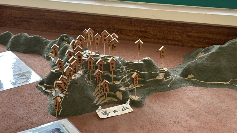 A three-dimensional terrain model showing Azuchi Castle's enclosures and building locations with labeled markers, giving a bird's-eye view of the mountaintop castle layout.
