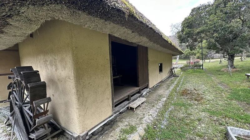 The exterior of the Former Miyaji Residence, with its thatched roof and earthen walls, and a wooden waterwheel set beside the building.