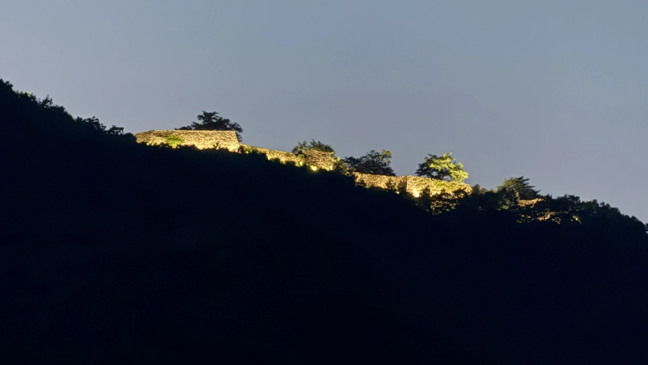 The illuminated stone walls of Takeda Castle Ruins standing out against the dark mountain ridge at night.