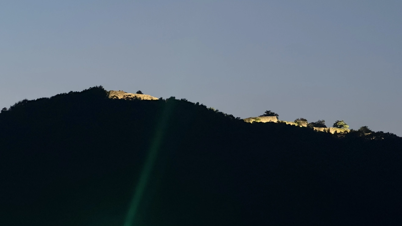 Takeda Castle Ruins lit up in patches along a dark mountain ridge, showing the outline of the site stretching along the ridgeline.