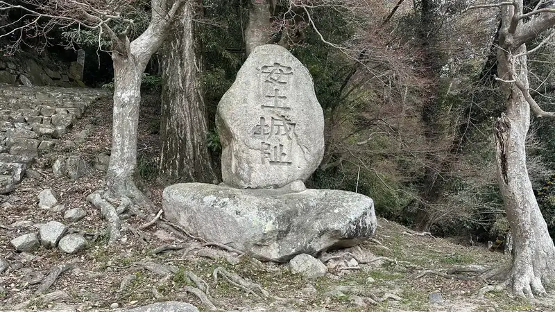 A stone marker reading 'Azuchi Castle Ruins' standing at Dodobashiguchi, with stone walls and the climbing path continuing up the slope behind it.