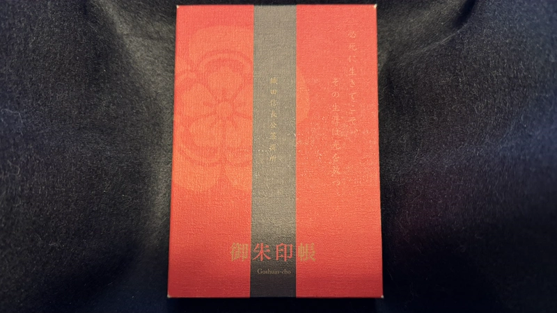 黒い布地の上に置かれた赤色と黒色の御朱印帳。「織田信長公菩提所」や「御朱印帳」の金文字に加え、表面に織田木瓜紋がうっすらとデザインされている。