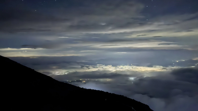 星空と街明かりを背に夜間の登山道を進む登山者