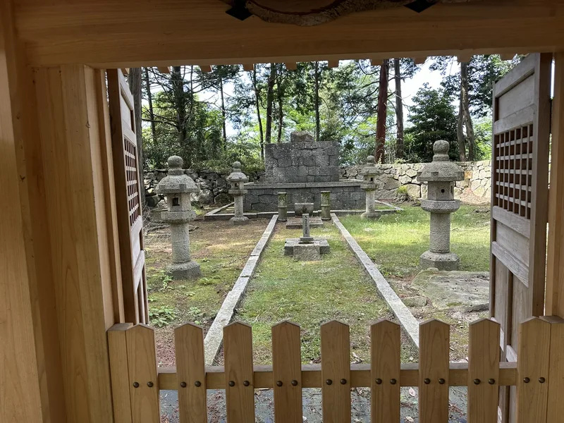 Nobunaga's Mausoleum viewed through a wooden gate. A stone burial site sits beyond a stone lantern.