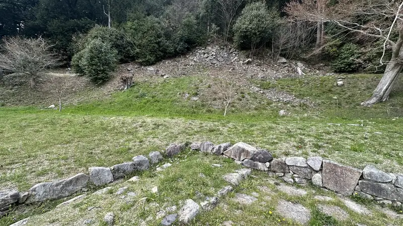 A view across the upper western enclosure, with a low row of stones in the foreground and a slope and stone wall beyond.