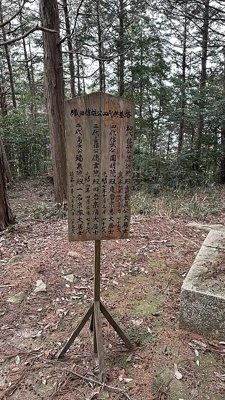 A wooden signboard in the woods explaining the location and background of the traditional Oda Nobukatsu mansion site.