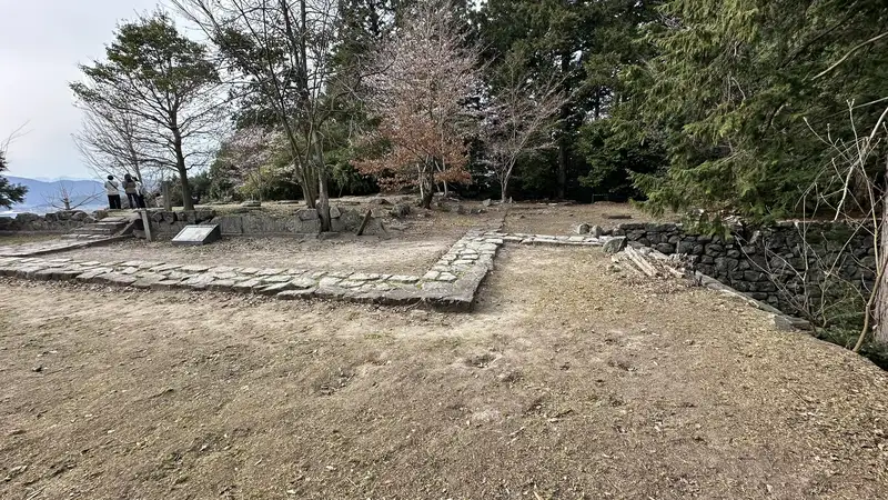 Foundation stones and stone rows remaining at the site of Sōken-ji's former main hall, with a view toward Lake Biwa through the trees.