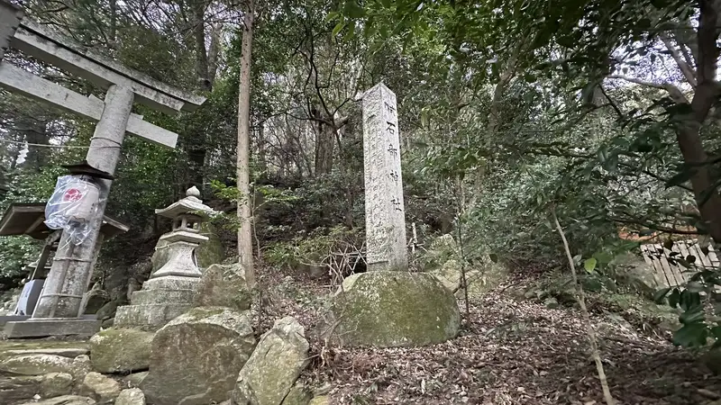 A stone marker for Ishibe Shrine standing beside a torii gate near Dodobashiguchi, surrounded by stone lanterns and trees.
