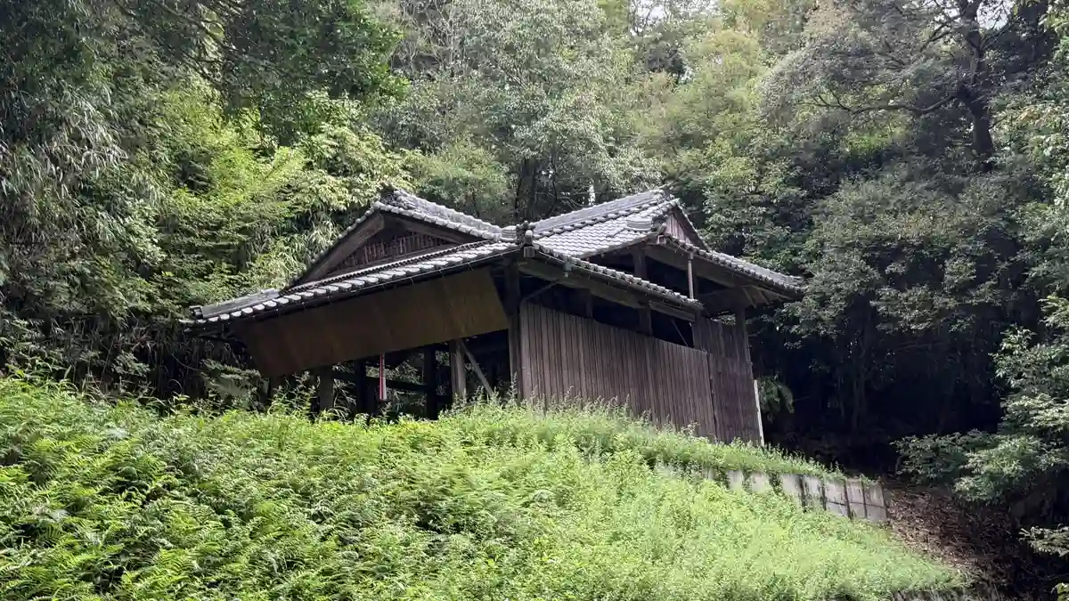 緑の斜面越しに見た、木々に囲まれた社殿の覆屋。屋根と木製の壁がのぞきます。