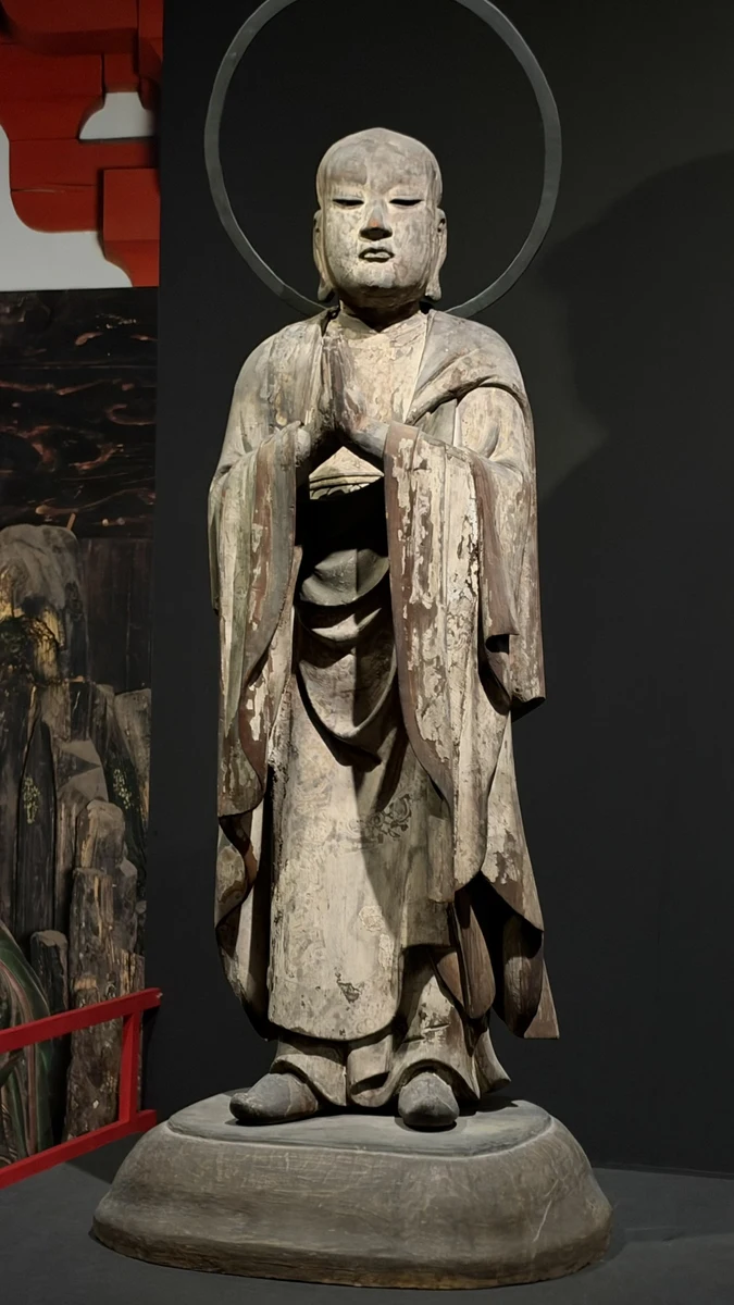 Ananda standing with joined hands, long robes and a circular halo